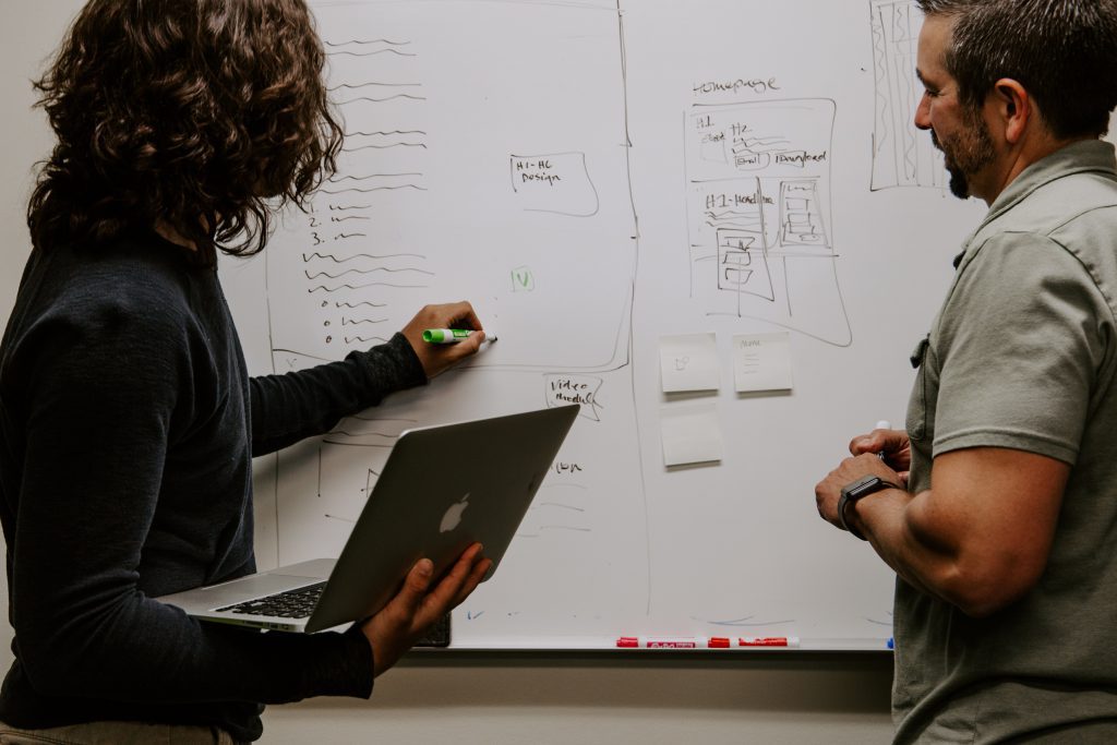 Two employees mentioning something on a Whiteboard holding a laptop in one hand